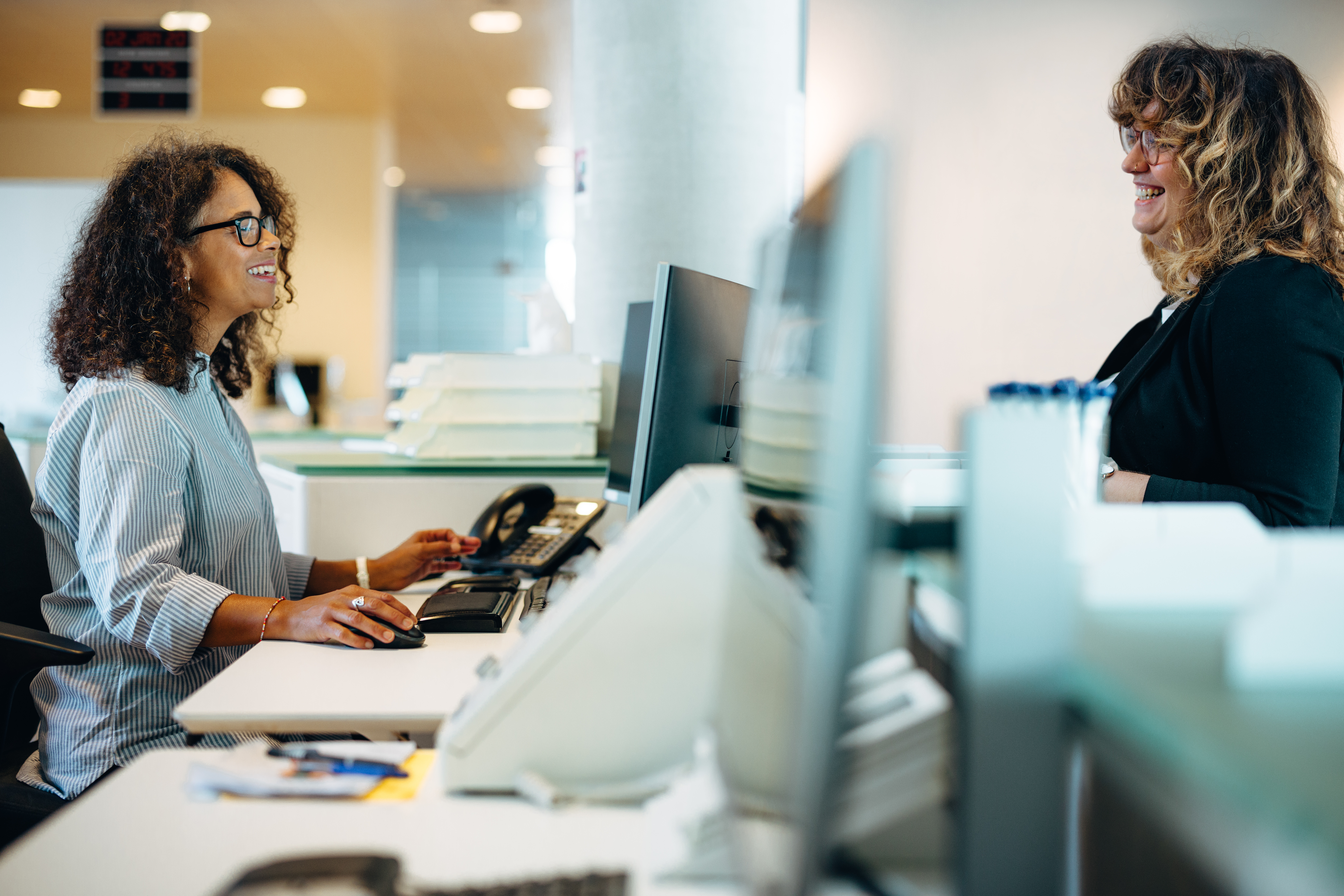 Smiling,Administrator,Talking,With,A,Woman,Standing,At,Reception,Desk. Smiling,Administrator,Talking,With,A,Woman,Standing,At,Reception,Desk.